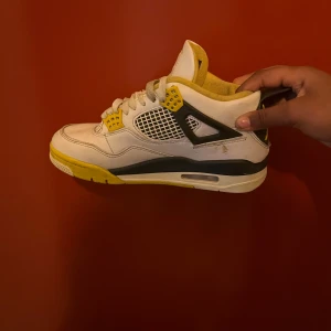 Nike Air Jordan 4 Retro vit/gul - Säljer ett par Nike Air Jordan 4 Retro sneakers i vitt med gula och svarta detaljer. Skorna har meshpaneler på sidorna, snörning och en bekväm platt sula. Perfekta för dig som gillar klassiska och färgstarka sneakers med ikonisk design.