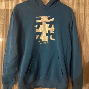 Blå hoodie från WARP med tryck - Säljer en blå hoodie från WARP med stort vitt grafiskt tryck på bröstet. Tröjan har huva och en klassisk känguruficka framtill. Perfekt för dig som gillar streetwear och vill ha en skön och snygg stil.