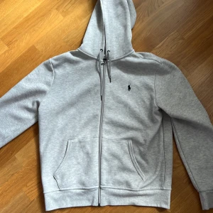 Grå hoodie från Polo Ralph Lauren - Snygg ljusgrå hoodie från Polo Ralph Lauren med dragkedja och klassisk logga på bröstet. Perfekt för chill dagar. Nypris 1200kr, mitt pris 600! 