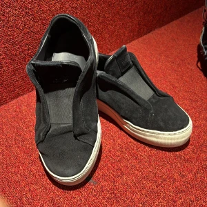 Svarta slip-on sneakers från Axel Arigato - Svarta slip-on sneakers från Axel Arigato i mocka med vit platt sula. Skorna har minimalistisk design utan snörning och diskret logga på plösen. Perfekta för en clean och stilren look.