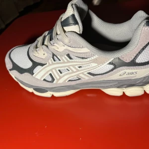 Asics Gel – Bekväma & Lätta Löparskor i Toppskick - Snygga Asics GEL sneakers i en mix av grått, beige och vitt. Skorna har meshpaneler för extra andningsförmåga, detaljer i mocka och syntet samt en chunky sula. Klassisk Asics-logga på sidan och broderad GEL-detalj bak. Perfekta för dig som gillar sportig och trendig stil.