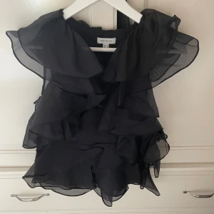 Svart volangblus från H&M Edition XS - Supersnygg svart blus från H&M Edition i storlek XS. Blusen är ärmlös och har flera lager av tunna, transparenta volanger som ger en cool och luftig känsla. Perfekt för dig som gillar statement-plagg med mycket rörelse och volym. 