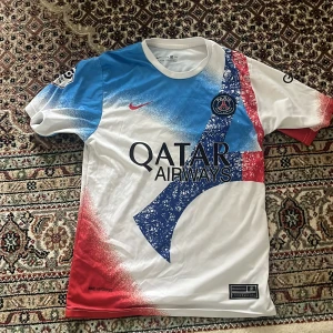 PSG Messi 30 Nike fotbollströja - Säljer en PSG fotbollströja med Messi 30 på ryggen. Tröjan är från Nike och har Dri-Fit material. Färgerna är vit, blå och röd med coolt grafiskt mönster och PSG-logga på bröstet. Qatar Airways och GOAT-loggor på framsida och ärm. Perfekt för fans!