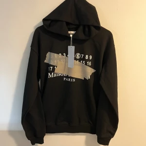 Maison Margiela Hoodie - Exklusiv hoodie från ikoniska Maison Margiela – känd för sina unika detaljer och tidlösa design. Tillverkad i högkvalitativ bomull som både känns lyxig och håller formen över tid. Minimalistisk men med den karakteristiska Margiela-touchen, perfekt för dig som vill ha något som sticker ut utan att bli för “loud”.  📏Storlek: S [ Längd = 63cm | Axelbredd = 55cm] 📦 Skick: [ Nyskick med Prislapp och Maison Margiela Paris påse]  Skriv gärna vid andra funderingar😊
