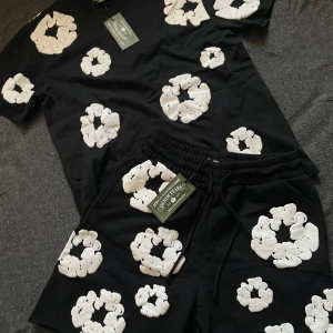 Säljer en Set från Denim Tears - Svart t-shirt från Denim Tears med stora vita blommor tryckta över hela plagget. T-shirten har rund hals och korta ärmar. Perfekt för dig som vill sticka ut med ett unikt och trendigt plagg.     Pris kan diskuteras 