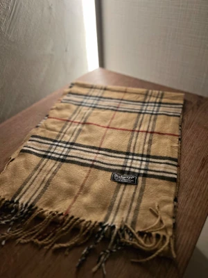 Rutig halsduk från Burberrys i ull - Klassisk rutig halsduk från Burberrys i 100% ull. Halsduken har fransar i ändarna och den ikoniska Burberrys-etiketten. Perfekt accessoar för att lyfta din outfit. Anledningen att de stör ”burberrys” är för att de är en äldre modell.