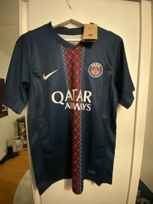 PSG Dembele #10 fotbollströja Nike - Säljer en Paris Saint-Germain fotbollströja med Ousmane Dembélé och nummer 10 på ryggen. Tröjan är marinblå med rött och vitt geometriskt mönster längs mitten, PSG-logga på bröstet och Qatar Airways som sponsor. Tillverkad i lätt polyester med korta ärmar.