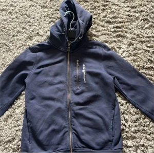 Blå hoodie från Sail Racing - Mörkblå hoodie från Sail Racing med dragkedja framtill och bröstficka med logga. Tröjan har huva och långa ärmar, perfekt för kyliga dagar. Snygg och enkel design som passar till det mesta.