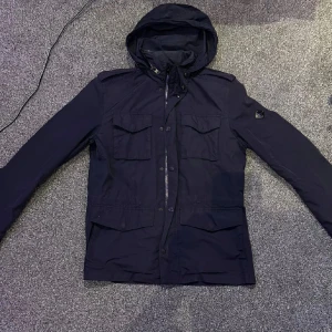 Mörkblå fieldjacka med infällbar luva från J.Lindeberg - Säljer en mörkblå jacka från J.Lindeberg med huva och flera fickor framtill. Jackan har dragkedja och knappar, samt en snygg logotyp på ärmen. Perfekt för vår och höst. Luvan går att dra in och ha en look utan luva
