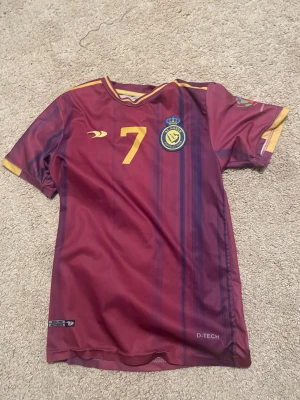 Al Nassr Ronaldo #7 vinröd matchtröja - Säljer en vinröd Al Nassr fotbollströja med gula detaljer och nummer 7, samt Ronaldo tryckt på ryggen. Tröjan har korta ärmar, tunna blå ränder och klubbmärke på bröstet. Materialet är lätt och andas, perfekt för träning eller match.