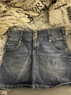 Blå jeanskjol med nitar och brodyr - Snygg blå jeanskjol med tre knappar framtill, klassiska fickor och coola nitar på myntfickan. Baksidan har broderade detaljer i färg och små nitar på fickorna. Kjolen har en kort modell och bred linning, perfekt för en trendig look.