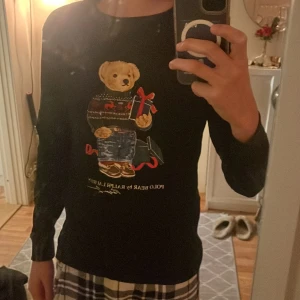 Svart Polo Bear tröja från Ralph Lauren - Svart långärmad tröja från Ralph Lauren med ikoniskt Polo Bear-tryck framtill. Björnen har stickad tröja, jeans och håller ett presentpaket med röd rosett. Tröjan har rund hals, perfekt för dig som gillar klassisk men lekfull stil.