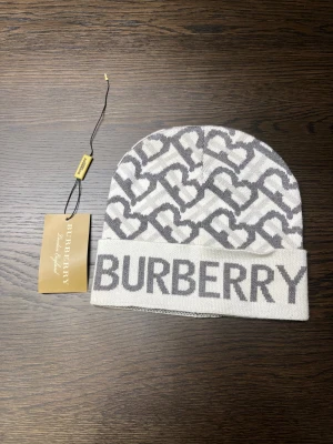 Burberry mössa med hjärtmönster - Snygg mössa från Burberry i ljusgrått och vitt med ett coolt hjärtmönster och stor logga framtill. Mjuk och skön stickad kvalitet som håller dig varm och ger en lyxig vibe. Perfekt accessoar för att lyfta din stil under kyliga dagar.