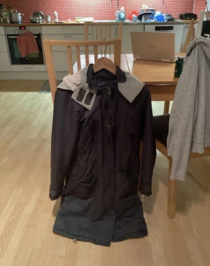 Svart parkajacka med teddyfoder - Svart parkajacka från Courage in Action med mjukt vitt teddyfoder på insidan och huvan. Jackan har dragkedja framtill, stora fickor och justerbar nederkant med snörning. Perfekt för kalla dagar och har en stilren, lång siluett.