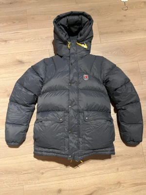 Fjällräven Expedition dunjacka mörkgrå - Säljer en mörkgrå Fjällräven Expedition Series dunjacka med huva och gula snören. Jackan har två stora fickor med knappar, dragkedja och tryckknappar framtill. Klassisk Fjällräven-logga på bröstet och quiltad design för extra värme. Helt ny skick!