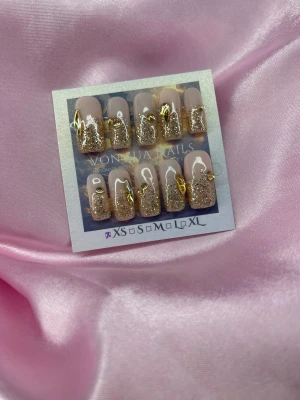 Glam(XS);Medium Square;Curved - Säljer ett set med handgjorda press-on naglar i nude och guld. Naglarna har en nude bas med glitter och skimrande gulddekorationer. Formen är medium square och de finns i XS: 14 mm, 11 mm, 11,5 mm, 10 mm, 8 mm. Perfekt för dig som vill ha extra glam på fingertopparna.