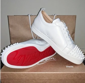 Vita Christian Louboutin sneakers äkta - Säljer ett par vita sneakers från Christian Louboutin med ikonisk röd sula och coola nitar framtill. Skorna har snörning och är tillverkade i slätt skinn för en lyxig känsla. Perfekt för dig som vill sticka ut med en edgy och exklusiv stil. Finns äkthets bevis