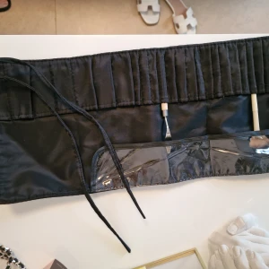 Svart sminkbälte med snörning - Svart sminkbälte med flera fack för penslar och tillbehör. Bältet har snörning för att knytas runt midjan och transparent plastficka för enkel översikt. Tillverkat i syntetmaterial med slät yta och stilren design, perfekt för dig som gillar att hålla ordning på sminket.