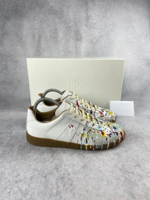 Maison Margiela dojor med färgstänk 🎨 👟  - Maison Margiela sneakers i vitt skinn med coola färgstänk i rött, blått, gult och grönt. Skorna har snörning, rund tå och en klassisk brun gummisula. Perfekt för dig som gillar unik design och vill sticka ut med dina skor. De är även helt nya köpt från hemsidan