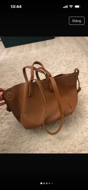 Brun handväska i skinn med axelrem - Snygg brun handväska i skinn med både handtag och axelrem. Väskan har en stilren design med magnetknapp och rymligt fack. Perfekt för dig som vill ha en klassisk och praktisk väska till vardagen.