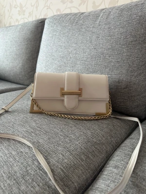 Vit axelväska från Aldo med kedja - Snygg vit axelväska från Aldo med stilrena gulddetaljer och kedja. Väskan har en rektangulär form, magnetlås och justerbar axelrem. Materialet är slätt syntet och insidan är ljusbeige. Perfekt för dig som gillar minimalistisk design och vill ha en väska som sticker ut.