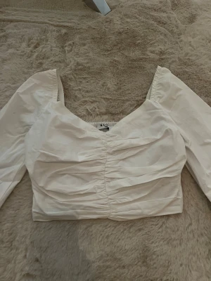 Vit croppad blus från NA-KD - Supersnygg vit blus från NA-KD i croppad modell med långa puffärmar och v-ringad hals. Toppen har snygga rynkade detaljer framtill. Helt oanvänd topp i storlek 32