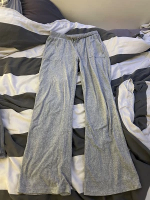 Grå mjukisbyxor från Juicy Couture - Grå mjukisbyxor från Juicy Couture med breda ben och snörning i midjan. Byxorna har fickor fram och bak, bakfickorna har lock med silverfärgade JC-knappar. Mjukt och skönt material, perfekt för chill dagar.