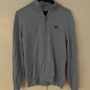 Ljusblå half zip-tröja från Lexington - Snygg ljusgrå tröja från Lexington med half zip och hög krage. Tröjan har lång ärm och liten logga på bröstet. Perfekt för dig som gillar stilrena och klassiska plagg med en modern touch.