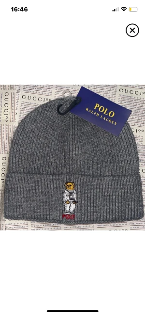 Grå ribbad mössa Polo Ralph Lauren - Snygg grå ribbstickad mössa från Polo Ralph Lauren med broderad nalle i astronautdräkt framtill. Perfekt accessoar för att hålla dig varm och ge din stil en cool touch. Klassisk uppvikt kant och mjukt material.