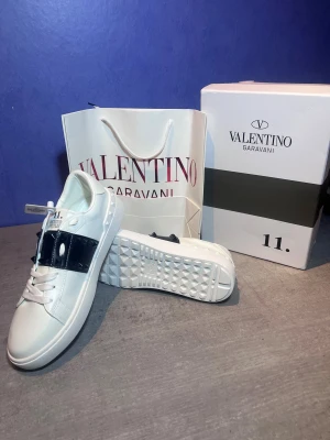Valentino Garavani Rockstud sneakers - Säljer ett par vita Valentino Garavani Rockstud Untitled sneakers med svart band och ikoniska nitar över vristen. Skorna har rund tå, platt sula och snörning. Tillverkade i lyxigt skinn med cool kontrast mellan vitt och svart. Perfekt för dig som gillar statement-detaljer.