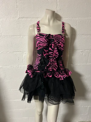 Zebramönstrad klänning med volanger S/M - Snygg klänning med rosa och svart zebramönster. Den har smala axelband och volanger framtill. Kjolen är svart med spetsdetaljer och ger en lekfull look. Helt ny. Storlek S/M.