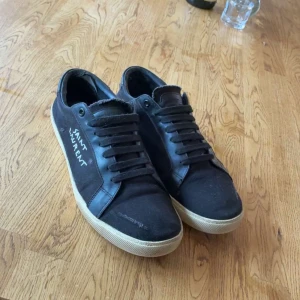Svarta Saint Laurent sneakers - Svarta sneakers från Saint Laurent med vit logga på sidan. Skorna har rund tå, platt beige sula och snörning framtill. Materialet är en mix av mocka och skinn vilket ger en lyxig känsla. Perfekt för dig som gillar stilrena och klassiska sneakers. Boxen och dustbag intlusive allt og följer med