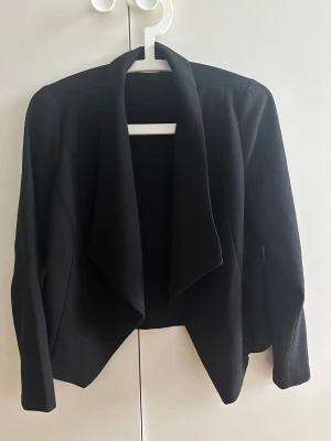 Svart kavaj med draperad front - Säljer en svart kavaj med snygg draperad front och lång ärm. Modellen är öppen framtill och har en lite loose passform som ger en chill men ändå uppklädd vibe. Perfekt till jeans eller kjol för en clean look