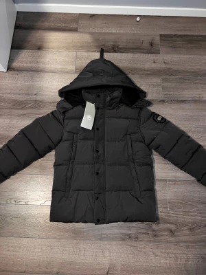 Svart pufferjacka från Canada Goose - Säljer en svart pufferjacka från Canada Goose med huva och diskret svart patch på ärmen. Jackan har knappar framtill, två stora fickor och quiltad design. Perfekt för kalla dagar och riktigt snygg till streetwear-looken.