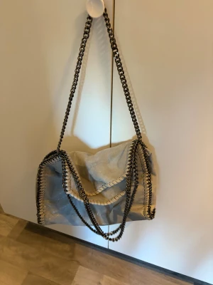 Grå axelväska med kedjedetaljer - Snygg grå axelväska från Stella McCartney med glansig mockayta och kraftiga kedjor som axelrem. Insidan har leopardmönstrat foder och en praktisk innerficka med dragkedja. Perfekt för dig som gillar coola detaljer och vill sticka ut.