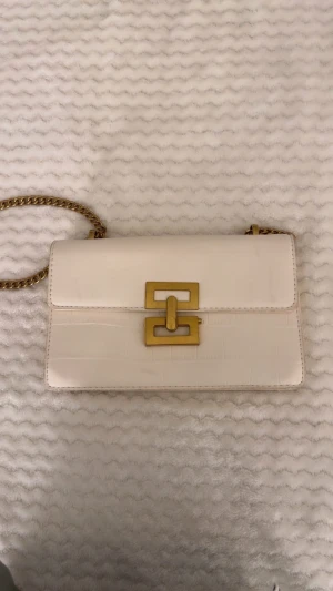 Vit väska, kedja och guldspänne - Snygg vit axelväska med croco-mönstrad yta och guldfärgat spänne framtill. Väskan har en kraftig guldfärgad kedja med axelrem i matchande vitt skinn. Insidan är svart och har två fack. Perfekt storlek för det viktigaste och ger en clean vibe.