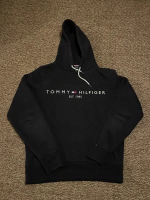 Svart hoodie Tommy Hilfiger - Svart hoodie från Tommy Hilfiger med stor logotyp och text 'EST. 1985' på bröstet. Tröjan har huva med vit snörning och en klassisk känguruficka framtill. Mjuk bomullskvalitet och långärmad modell, perfekt för chill dagar.