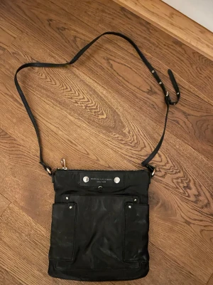 Svart axelväska från Marc by Marc Jacobs - Snygg svart axelväska från Marc by Marc Jacobs med justerbar rem och silverfärgade detaljer. Väskan har två praktiska fickor framtill och dragkedja upptill. Perfekt storlek för mobil, plånbok och andra essentials.