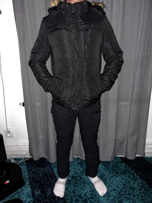 Svart pufferjacka med fuskpäls - Svart pufferjacka med huva och snygg fuskpälsdetalj. Jackan har dragkedja och knappar framtill, samt praktiska fickor på sidorna och bröstet. Materialet är glansigt syntet och jackan är quiltad för extra värme. Perfekt för kalla dagar och har en normal passform.