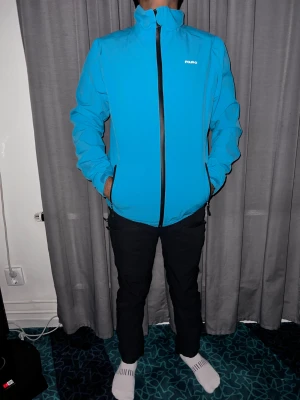 Blå vindjacka från FOUR-D, storlek S - Snygg blå vindjacka från FOUR-D med svart dragkedja och hög krage. Jackan är tillverkad i syntetmaterial som är både vattentätt och andas. Perfekt för dig som gillar sportig stil och vill ha en jacka som står emot väder och vind. Två sidofickor med dragkedja.