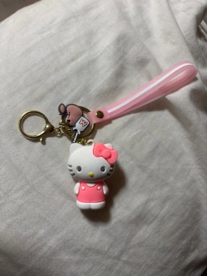 Hello Kitty nyckelring med rosa band - Supergullig Hello Kitty nyckelring med en söt figur i vitt och rosa, komplett med en rosa rosett och klänning. Nyckelringen har även ett mjukt rosa handledsband och en liten musdetalj. Perfekt accessoar för att piffa upp väskan eller nycklarna.