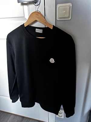 Svart sweatshirt från Moncler - Svart sweatshirt från Moncler med rund hals och lång ärm. Tröjan har ett stilrent Moncler-logo patch på bröstet och ribbade muddar vid ärmslut och nederkant. Perfekt för dig som gillar clean och exklusiv streetwear.