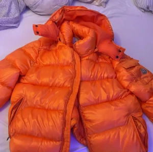 Orange dunjacka från Moncler - Säljer en fet orange dunjacka från Moncler med hög krage och huva. Jackan har Moncler-logga på ärmen, dragkedja och tryckknappar framtill samt flera fickor. Materialet är glansig polyester och dunfyllning, perfekt för kalla dagar. Taggar och etiketter sitter kvar. QR KOD och NFC!