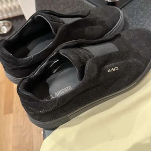 Arigato skor - Svarta skor från Axel Arigato i mocka med elastisk panel över vristen för enkel påtagning. Diskret logga i guld på sidan och på plösen. Snygg, minimalistisk design med rund tå och platt sula. Perfekt för dig som gillar stilrena och moderna skor. Påse tillkommer.