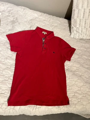 Burberry piké - Snygg röd pikétröja från Burberry i slim fit med klassisk krage och korta ärmar. Ikonisk Burberry-rutig detalj vid knäppningen och liten broderad logga på bröstet. Tillverkad i bomull och perfekt för en clean och stilren look.