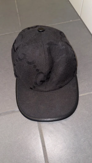 Svart keps från Gucci med GG-mönster - Snygg svart keps från Gucci med diskret GG-logomönster över hela kepsen. Justerbar rem bak i svart skinn och klassisk böjd skärm. Materialet är robust bomull med detaljer i skinn. Kommer i original Gucci-box.