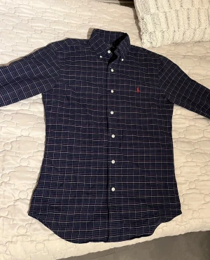 Rutig blå skjorta Polo Ralph Lauren - Säljer en slim fit rutig skjorta från Polo Ralph Lauren i blått med inslag av rött, grönt och vitt. Skjortan har klassisk krage, knappar framtill och lång ärm. Ikoniska röda loggan broderad på bröstet. Perfekt för dig som gillar stilren och tidlös look.
