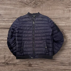 Mörkblå pufferjacka Polo Ralph Lauren - Snygg mörkblå pufferjacka från Polo Ralph Lauren med broderad logga på ärmen. Jackan har ribbad krage, dragkedja framtill och två fickor med knappstängning. Quiltad design och klassisk bomberform, perfekt för kyliga dagar.