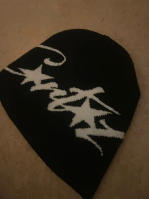 Corteiz Mössa - Svart mössa från Corteiz med vit, stickad graffiti-logga och stjärndetaljer på framsidan. Mjuk och skön passform, perfekt för dig som gillar streetwear och vill sticka ut med en unik accessoar. Materialet är stickat och designen är riktigt clean.