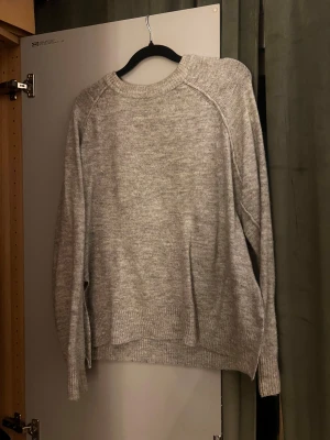 Grå stickad tröja med rund hals - Mysig grå stickad tröja med rund hals och långa ärmar. Tröjan har en enkel design med ribbade muddar vid ärmslut och nederkant. 